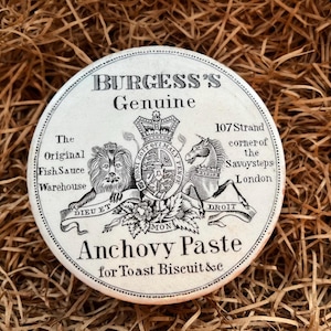 May include: A white ceramic jar lid with black lettering that reads "BURGESS'S Genuine The Original Fish Sauce Warehouse 107 Strand corner of the Savoy steps London Anchovy Paste for Toast Biscuit &c". The lid features a black and white illustration of a lion, a unicorn, and a coat of arms.