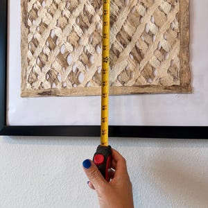 May include: A framed artwork with a woven, textured design in shades of beige and brown. A yellow measuring tape shows the artwork is approximately 30 inches in length. The artwork is framed in a black frame and mounted on a white wall.