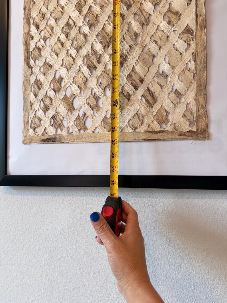 May include: A framed artwork with a woven, textured design in shades of beige and brown. A yellow measuring tape shows the artwork is approximately 30 inches in length. The artwork is framed in a black frame and mounted on a white wall.