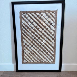 May include: Framed artwork featuring a textured, brown and white diagonal striped design. The artwork is set against a white background within a black frame. The piece is displayed on a wooden floor.