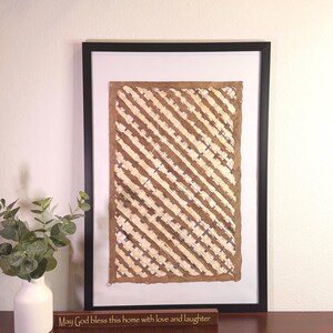 May include: Framed artwork with a brown and cream diagonal striped pattern. A small wooden sign below the artwork reads "May God bless this home with love and laughter." A white vase with greenery is on the left.
