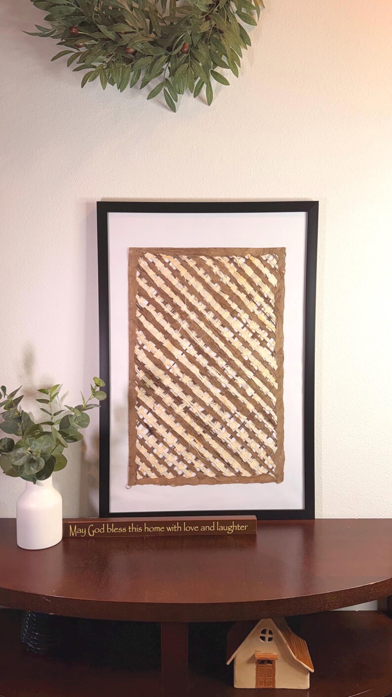 May include: Framed artwork with a brown and cream diagonal striped pattern. A small wooden sign below the artwork reads "May God bless this home with love and laughter." A white vase with greenery is on the left.