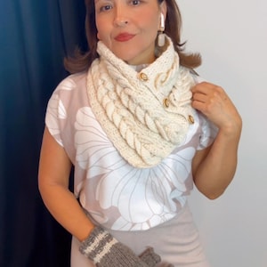 May include: Cream-colored knitted cowl neck warmer with a braided design and wooden buttons. The image also shows gray fingerless gloves with white stripes and a floral print top. A cozy winter accessory.