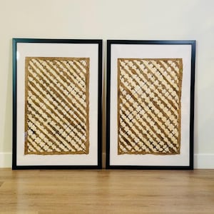 May include: Two framed artworks. Each piece features a textured, woven design in shades of beige and brown, set against a white background. The artwork is encased in a black frame, creating a striking contrast. The overall aesthetic is natural and earthy.