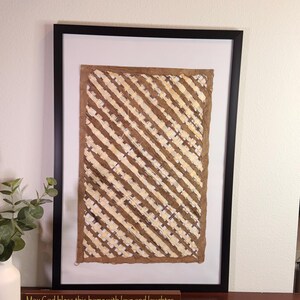 May include: Framed artwork with a brown and cream diagonal pattern. The artwork is in a black frame and is displayed on a dark brown table. A wooden sign below the artwork reads "May God bless this home with love and laughter."