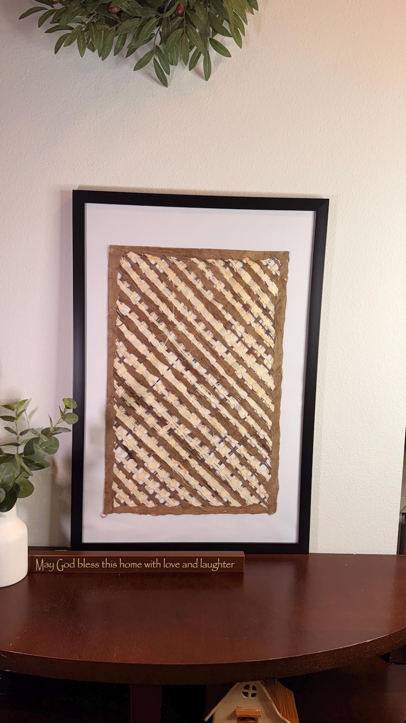 May include: Framed artwork with a brown and cream diagonal pattern. The artwork is in a black frame and is displayed on a dark brown table. A wooden sign below the artwork reads "May God bless this home with love and laughter."