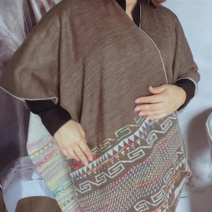 May include: A brown poncho with a geometric patterned border in shades of red, yellow, green, and white. The poncho has a white trim and is worn over a black long-sleeved shirt. The poncho is made of a soft, woven material.