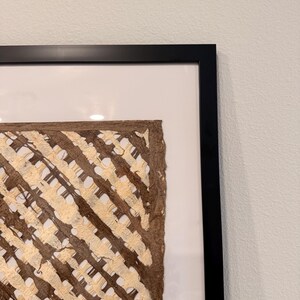 May include: A framed artwork featuring a textured, woven design in shades of brown and beige. The artwork is encased in a black frame with a white matting, creating a striking contrast. The woven pattern adds a tactile and visual element to the piece.