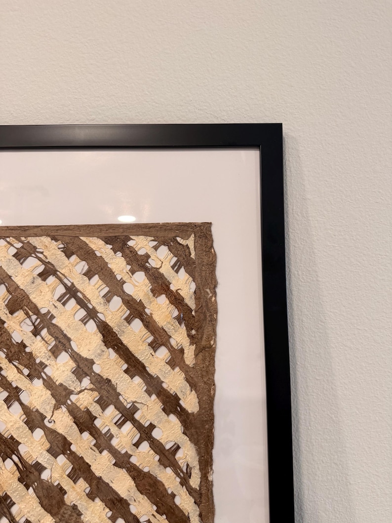 May include: A framed artwork featuring a textured, woven design in shades of brown and beige. The artwork is encased in a black frame with a white matting, creating a striking contrast. The woven pattern adds a tactile and visual element to the piece.