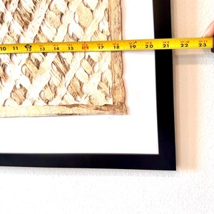 May include: A framed artwork featuring a textured, woven design in shades of beige and brown. The artwork is framed in a black frame and measures approximately 23 inches in length.