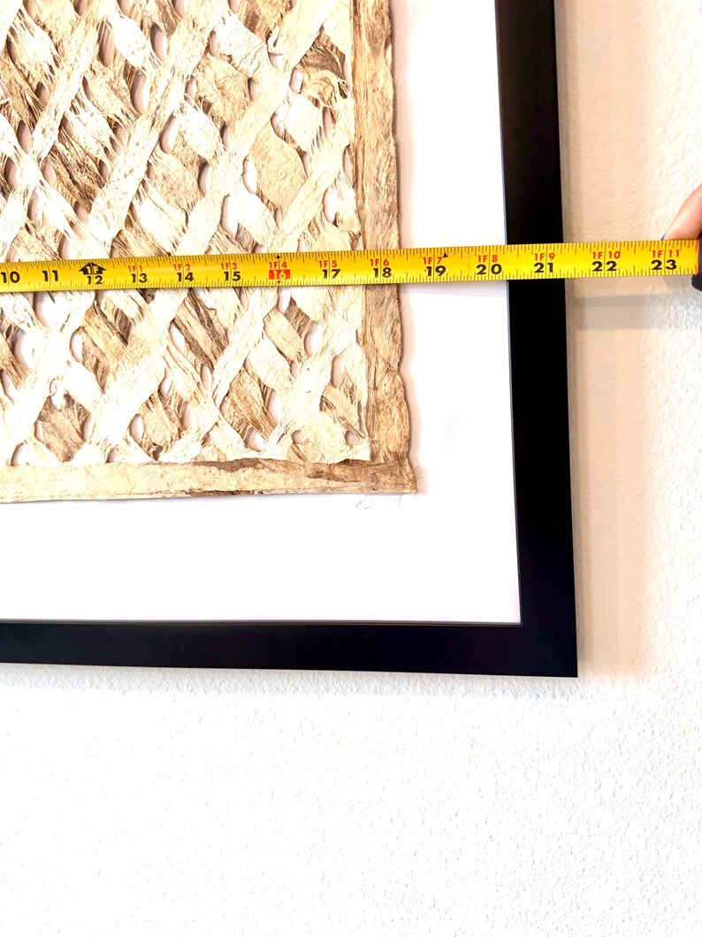 May include: A framed artwork featuring a textured, woven design in shades of beige and brown. The artwork is framed in a black frame and measures approximately 23 inches in length.
