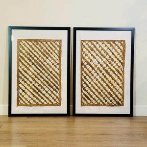May include: Two framed artworks. Each piece features a textured, woven design in shades of beige and brown, set against a white background. The artwork is encased in a black frame, creating a striking contrast. The overall aesthetic is natural and earthy.
