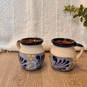 May include: Two handmade ceramic mugs with a beige base, a dark blue rim and handle, and a decorative pattern in blue, white, and yellow. The mugs are on a wooden surface.