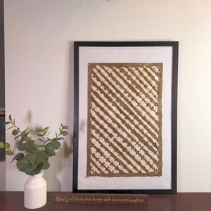 May include: A framed artwork with a black frame, featuring a brown and white diagonal striped pattern. The artwork is displayed on a dark brown table, next to a white vase with green leaves and a wooden sign that reads "May God bless this home with love and laughter."