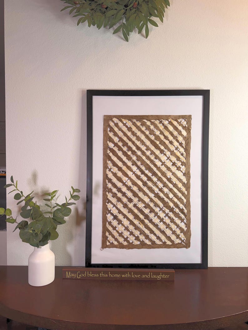 May include: A framed artwork with a black frame, featuring a brown and white diagonal striped pattern. The artwork is displayed on a dark brown table, next to a white vase with green leaves and a wooden sign that reads "May God bless this home with love and laughter."