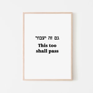 May include: A framed print with the phrase "This too shall pass" in English and Hebrew script. The text is in black against a white background, framed by a light wood frame. The artwork is minimalist and suitable for home decor.