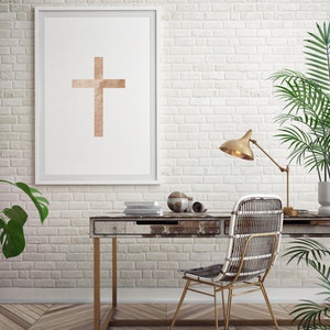 May include: A white framed print of a minimalist cross design in a light brown color hangs on a white brick wall. A wooden desk with a drawer sits in front of the print. A wicker chair with a gold metal frame sits at the desk. A gold lamp with a white shade sits on the desk. A potted plant sits on the floor next to the desk.