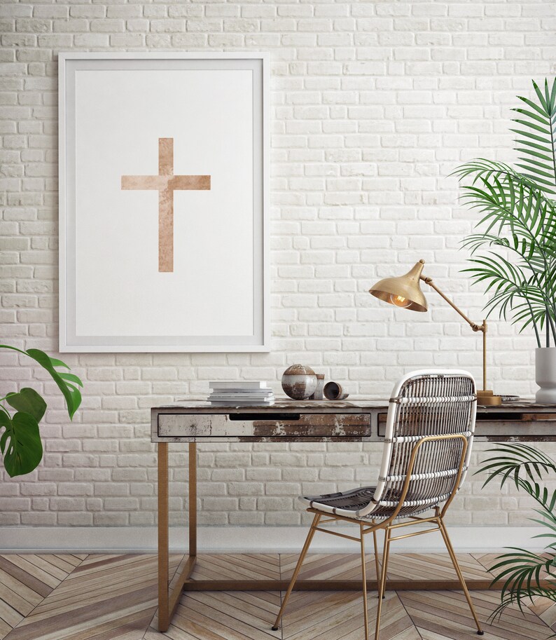 May include: A white framed print of a minimalist cross design in a light brown color hangs on a white brick wall. A wooden desk with a drawer sits in front of the print. A wicker chair with a gold metal frame sits at the desk. A gold lamp with a white shade sits on the desk. A potted plant sits on the floor next to the desk.