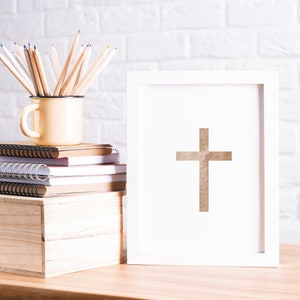 May include: A white picture frame with a brown cross inside. The frame is sitting on a wooden table next to a stack of notebooks and a yellow mug with colored pencils.
