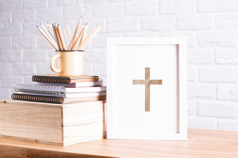 May include: A white picture frame with a brown cross inside. The frame is sitting on a wooden table next to a stack of notebooks and a yellow mug with colored pencils.