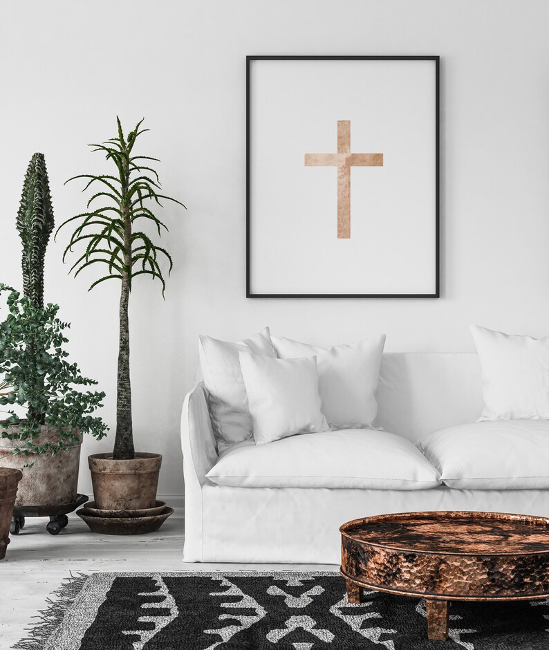 May include: A white couch with two pillows sits in front of a white wall with a framed print of a brown cross. A black and white patterned rug is on the floor in front of the couch. A round, copper-colored coffee table sits in front of the couch.