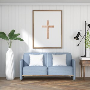 May include: A light blue sofa with two white pillows in front of a white wall with a framed picture of a brown cross. A black lamp with a white shade is on a wooden table to the right of the sofa.