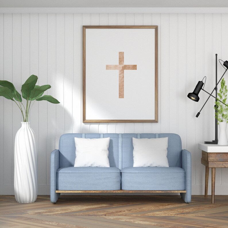 May include: A light blue sofa with two white pillows in front of a white wall with a framed picture of a brown cross. A black lamp with a white shade is on a wooden table to the right of the sofa.