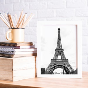 May include: A white framed black and white photograph of the Eiffel Tower in Paris, France. The photo is sitting on a wooden table with a stack of notebooks, a mug filled with colored pencils, and a wooden box.
