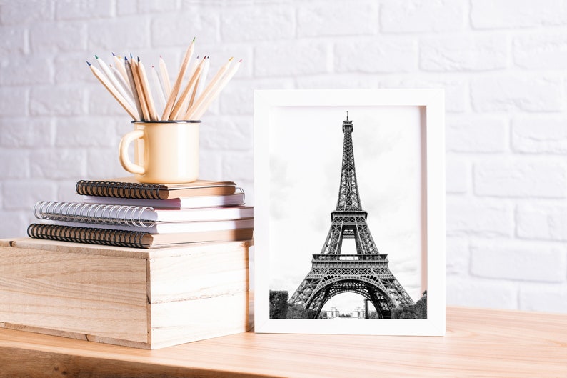 May include: A white framed black and white photograph of the Eiffel Tower in Paris, France. The photo is sitting on a wooden table with a stack of notebooks, a mug filled with colored pencils, and a wooden box.