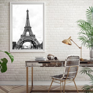May include: A black and white photograph of the Eiffel Tower in Paris, France, framed in a white frame and hanging on a white brick wall. A wooden desk with a gold metal frame sits in front of the wall with a wicker chair and a gold lamp.