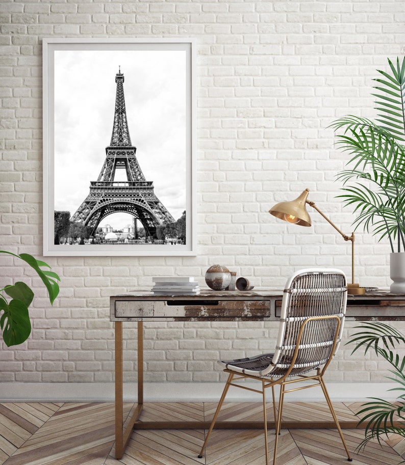 May include: A black and white photograph of the Eiffel Tower in Paris, France, framed in a white frame and hanging on a white brick wall. A wooden desk with a gold metal frame sits in front of the wall with a wicker chair and a gold lamp.