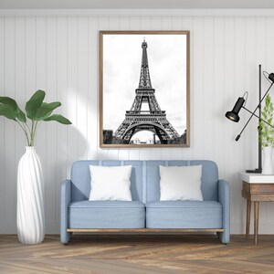 May include: A black and white photograph of the Eiffel Tower in Paris, France. The photo is framed in a light wood frame and is hanging on a white wall above a light blue sofa with white pillows.