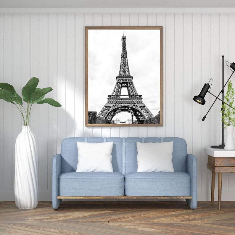 May include: A black and white photograph of the Eiffel Tower in Paris, France. The photo is framed in a light wood frame and is hanging on a white wall above a light blue sofa with white pillows.