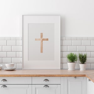 May include: A white framed print with a gold cross on a light gray background. The print is hanging on a white tiled wall above a white kitchen counter with wooden countertop and white cabinets.