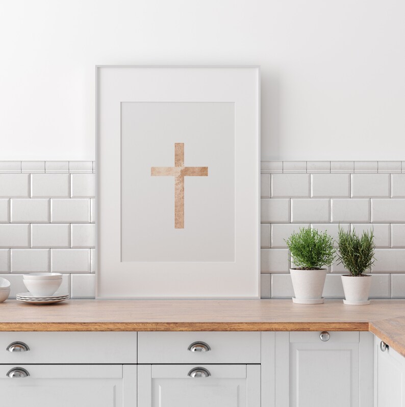 May include: A white framed print with a gold cross on a light gray background. The print is hanging on a white tiled wall above a white kitchen counter with wooden countertop and white cabinets.