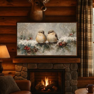 May include: A framed painting depicts two small birds perched on a snow-covered evergreen branch with red berries and pine cones. The artwork is displayed above a stone fireplace in a rustic, wood-paneled room.
