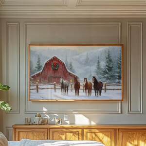 K&ouml;nnte beinhalten: Ein gerahmtes Gem&auml;lde zeigt eine Winterlandschaft mit einer roten Scheune, die mit einem Weihnachtskranz und Lichtern geschm&uuml;ckt ist. Vier Pferde stehen im Schnee vor der Scheune. Das Kunstwerk ist in einem Holzrahmen gefasst und h&auml;ngt &uuml;ber einem Holzschrank.