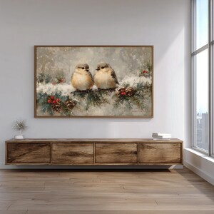 May include: A framed painting depicts two small birds perched on a snow-covered branch with pine needles, red berries, and pine cones. The artwork has a neutral color palette with a winter theme, suitable for home decor.