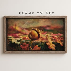 Puede incluir: Una obra de arte enmarcada que presenta una pintura detallada de una bellota con su gorro, descansando sobre un tocón de árbol cubierto de musgo. El fondo incluye hojas de otoño coloridas en tonos de rojo, naranja y verde. El texto "FRAME TV ART" se muestra en la parte superior.