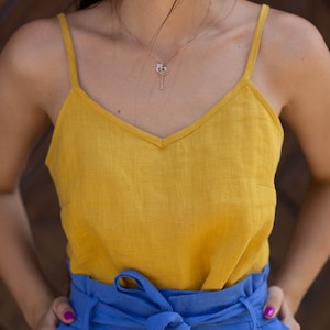 Puede incluir: Una camiseta sin mangas de color amarillo mostaza con cuello en V se combina con pantalones azules de cintura alta y pernera ancha. Los pantalones tienen un cinturón de tela atado con un lazo en la cintura. Se ve un delicado collar con colgante de llave.