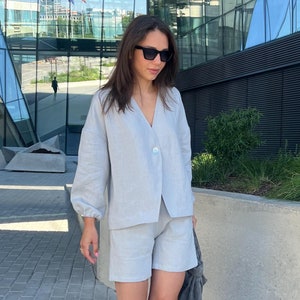 Puede incluir: Conjunto de shorts de lino gris claro, que consta de una camisa con botones y pantalones cortos. El atuendo se combina con sandalias azules y un bolso tote gris. La persona lleva gafas de sol y camina por una acera de ladrillos.