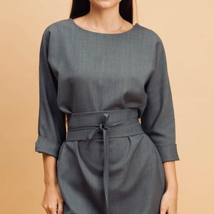 May include: A grey dress with a boat neck and three-quarter sleeves. The dress has a tie-waist detail and a knee-length skirt. The fabric has a subtle texture, and the overall style is simple and elegant.