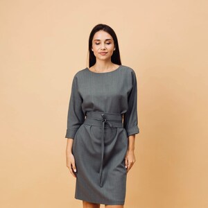 May include: A grey dress with a matching belt and three-quarter sleeves. The dress has a knee-length skirt and is paired with black ankle boots. The dress is made of a textured fabric.
