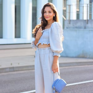K&ouml;nnte beinhalten: Hellblaues Crop-Top mit Puff&auml;rmeln und passender hochtaillierter Hose. Eine kleine, hellblaue Handtasche mit Quaste wird gehalten. Das Outfit ist aus leichtem Stoff gefertigt, perfekt f&uuml;r warmes Wetter.