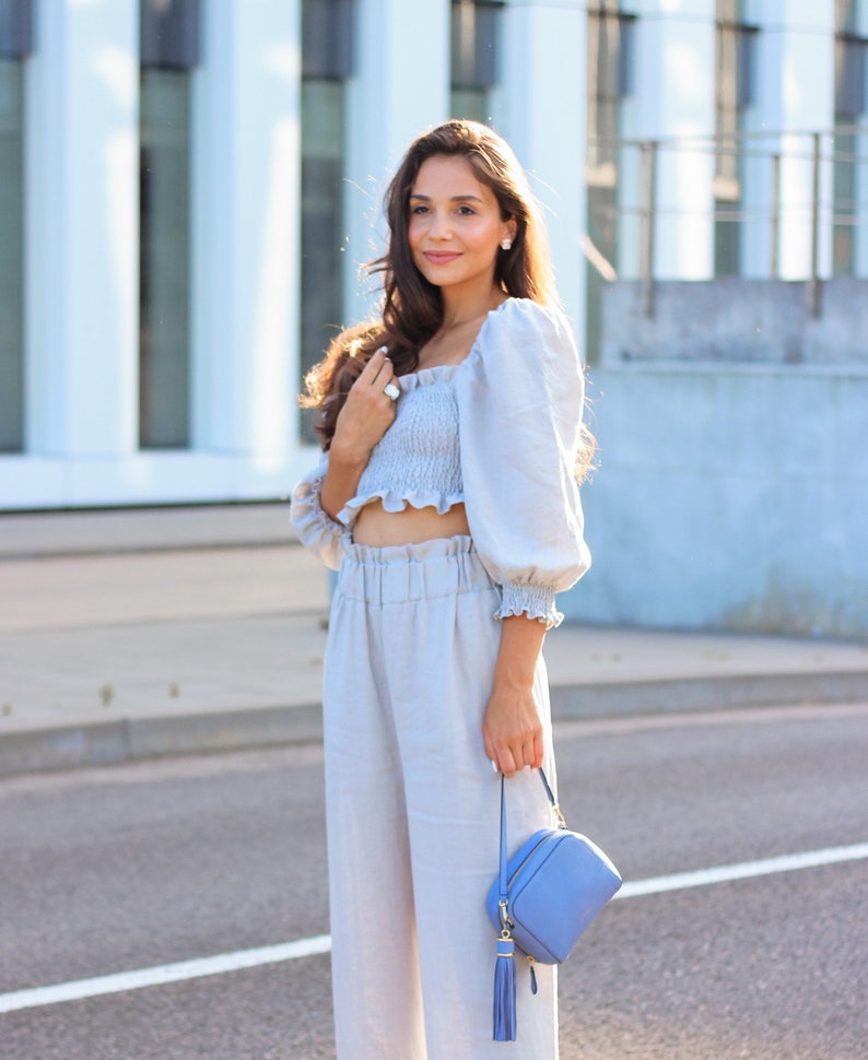 K&ouml;nnte beinhalten: Hellblaues Crop-Top mit Puff&auml;rmeln und passender hochtaillierter Hose. Eine kleine, hellblaue Handtasche mit Quaste wird gehalten. Das Outfit ist aus leichtem Stoff gefertigt, perfekt f&uuml;r warmes Wetter.