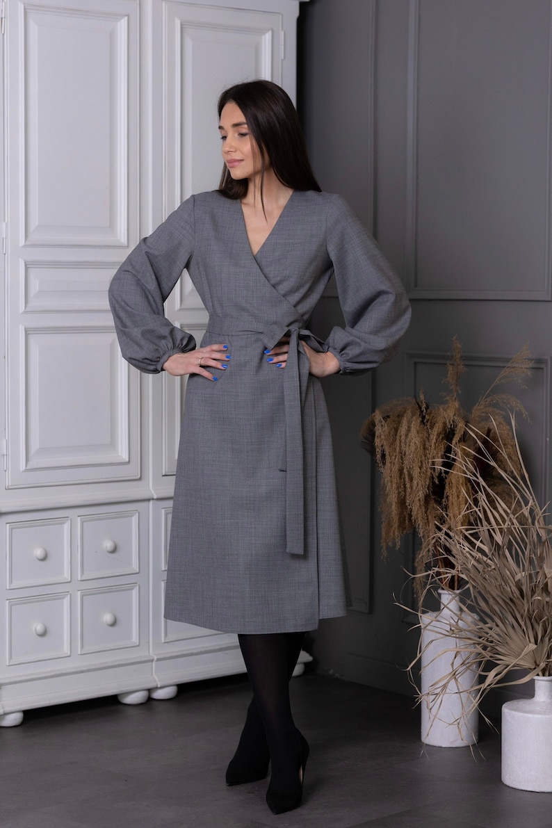 May include: A gray wrap dress with long sleeves and a matching belt. The dress has a V-neck and falls to the mid-calf. The model is wearing black tights and heels. The background includes a white cabinet and decorative plants.