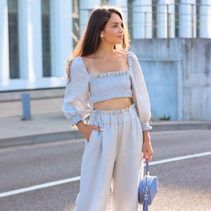 K&ouml;nnte beinhalten: Hellblaues Leinen-Ensemble, bestehend aus einem Crop-Top mit quadratischem Ausschnitt und langen, gerafften &Auml;rmeln. Passende hochtaillierte, weite Hose. Eine kleine, quadratische, hellblaue Handtasche mit Quaste.