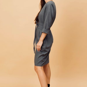 May include: A grey dress with a matching belt and rolled-up sleeves. The dress is knee-length and worn with black ankle boots. The model is in profile against a neutral background.