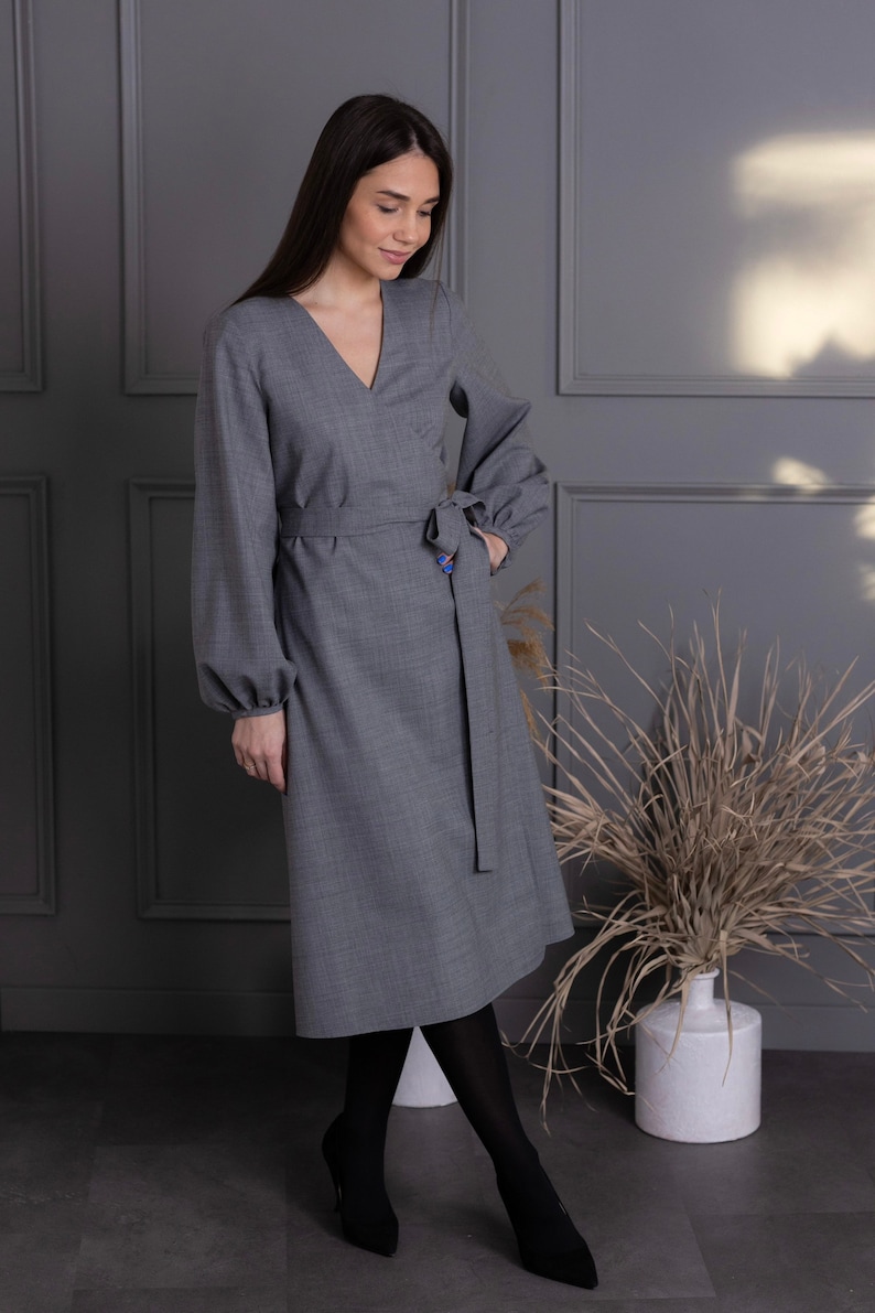 May include: A gray wrap dress with long sleeves and a V-neck. The dress features a matching belt that ties at the waist. The model is wearing black tights and black heels. The dress is knee-length and made of a textured fabric.