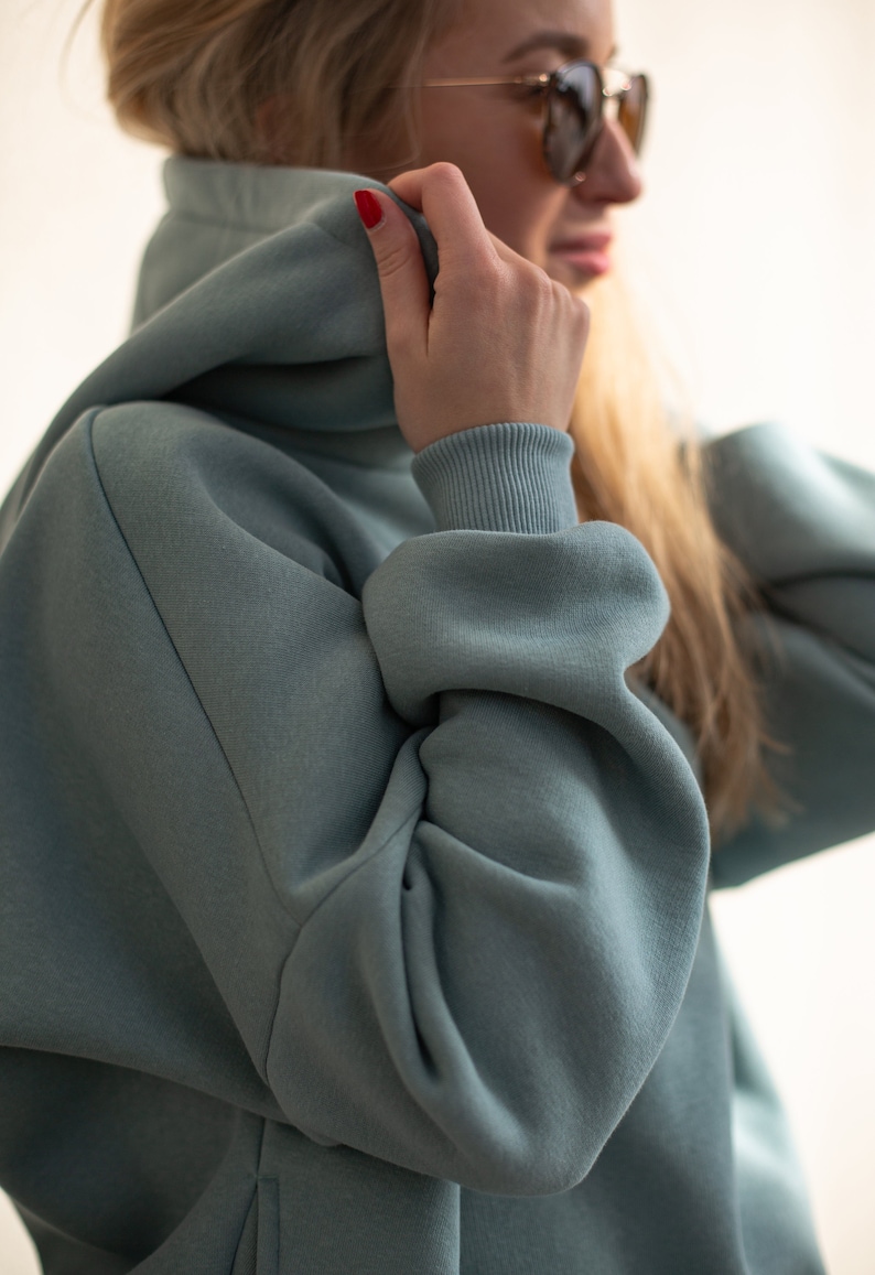 K&ouml;nnte beinhalten: Ein hellblaues Kapuzenpullover mit hohem Kragen und Kapuze. Die &Auml;rmel haben gerippte B&uuml;ndchen. Die Person auf dem Bild tr&auml;gt eine Sonnenbrille und hat blondes Haar. Der Kapuzenpullover ist aus einem weichen, dicken Material.